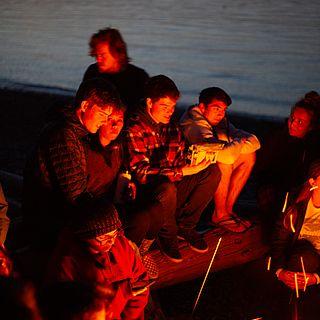 Several students sit around a campfire on the beach, their faces are orange with the glow from the fire.