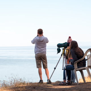 Three students stand on a bluff overlooking the ocean. One has binoculars and another a spotting scope.