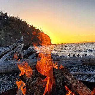A fire on the beach in the foreground, a glowing sunset over the ocean in the background.